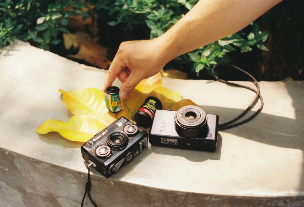 A hand arranges retro cameras and film on yellow leaves outdoors in autumn.